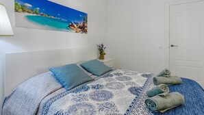 Ett sovrum med en säng, kuddar, en tavla som föreställer en strand, en vas med blommor och en dörr.