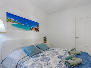 A bedroom with a bed, pillows, a painting of a beach, a vase with flowers, and a door.