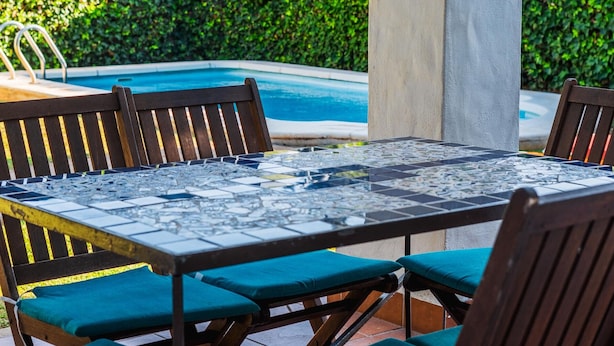 A poolside area with a tiled table and chairs, a pool, and a hedge in the background.