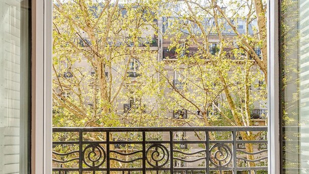 Un balcon orné d’une balustrade en fer forgé, une vue sur une rue bordée d’arbres et, au loin, un immeuble avec de nombreuses fenêtres.