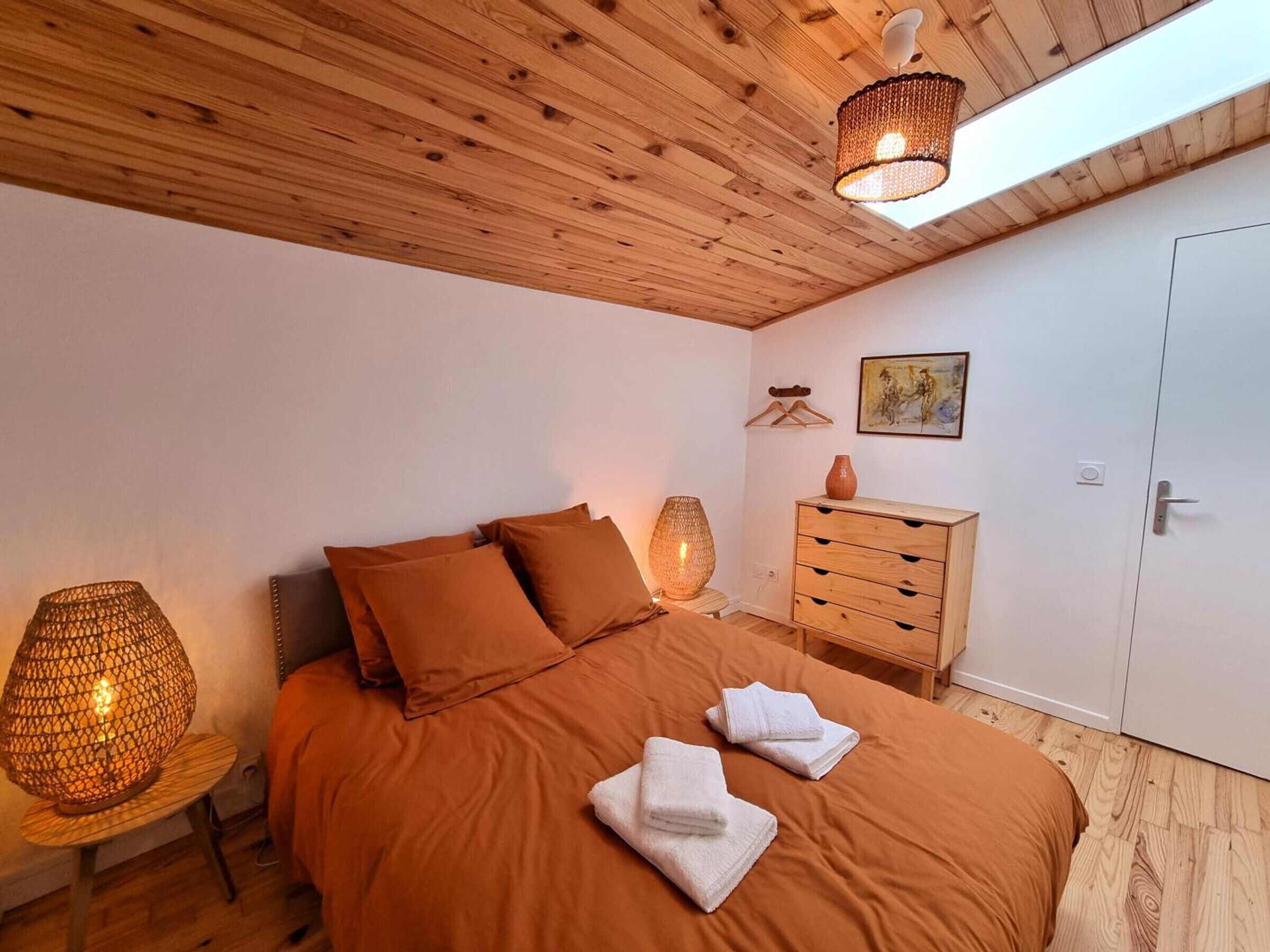 A bedroom with a wooden ceiling, a bed with brown bedding, a wooden dresser, and a wall-mounted painting.