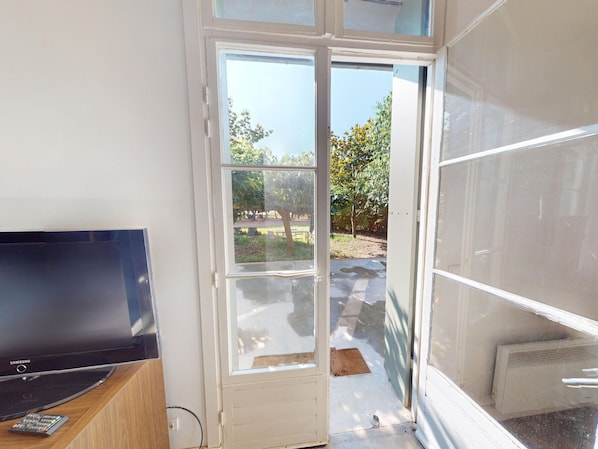Ein Zimmer mit Samsung Fernseher auf einem Holzboden, eine Glastür zur Terrasse mit Bank und Bäumen.