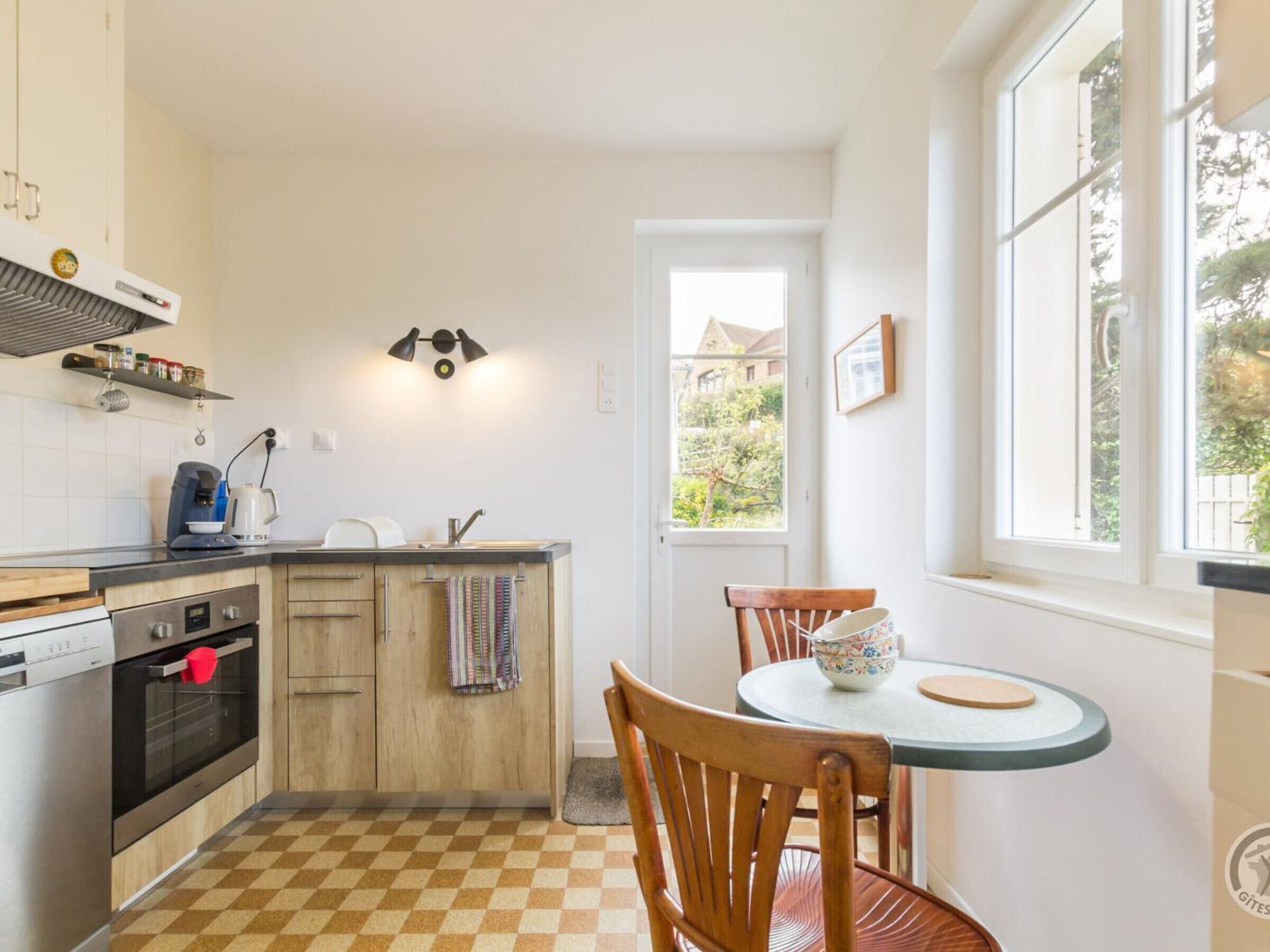 A compact kitchen with a checkered floor, wooden cabinets, a built-in oven, a coffee maker, and a window with a view of greenery.