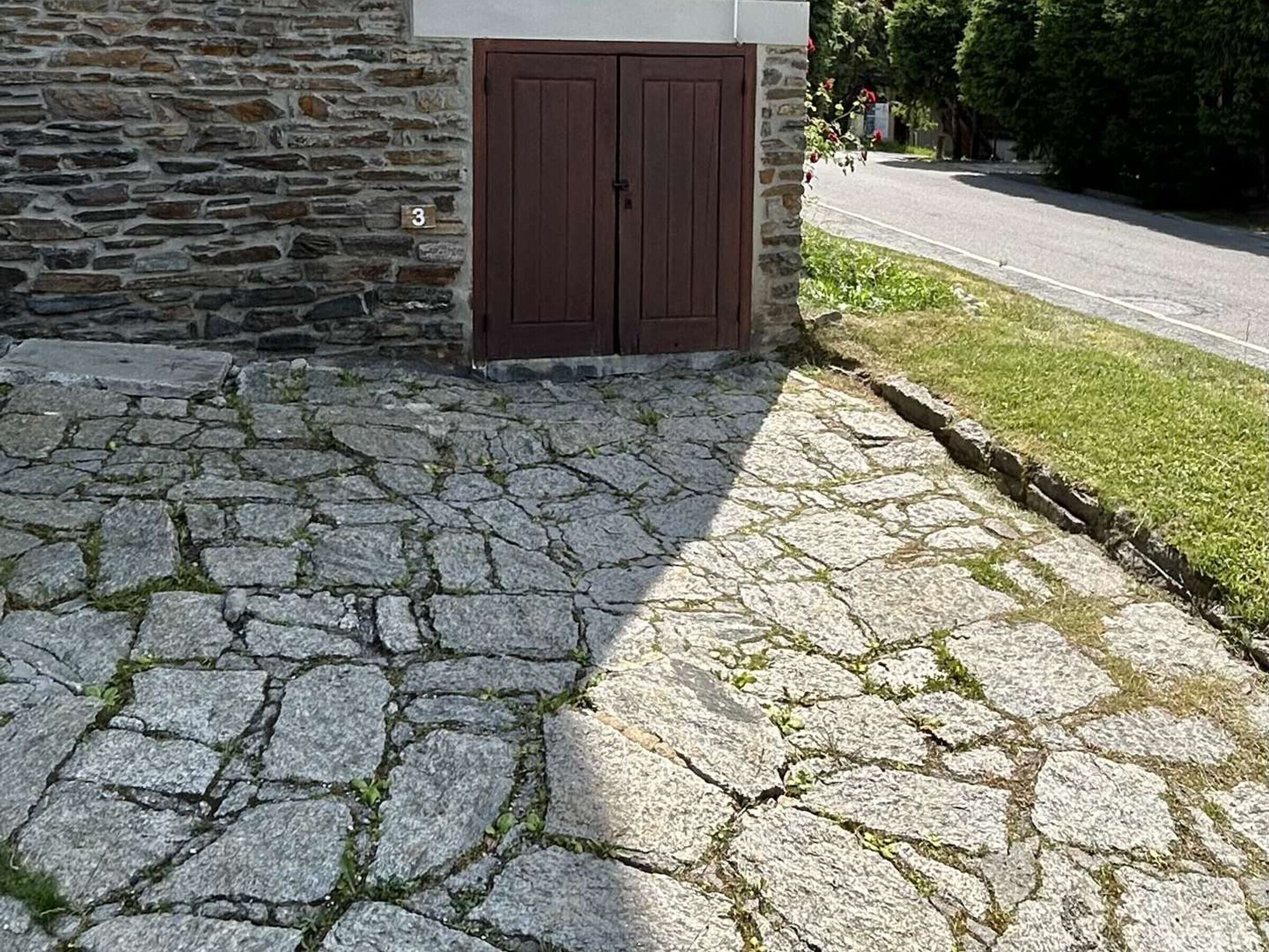 Un edificio de piedra con un doble portal de madera, un acceso adoquinado y una zona verde al lado de una carretera.