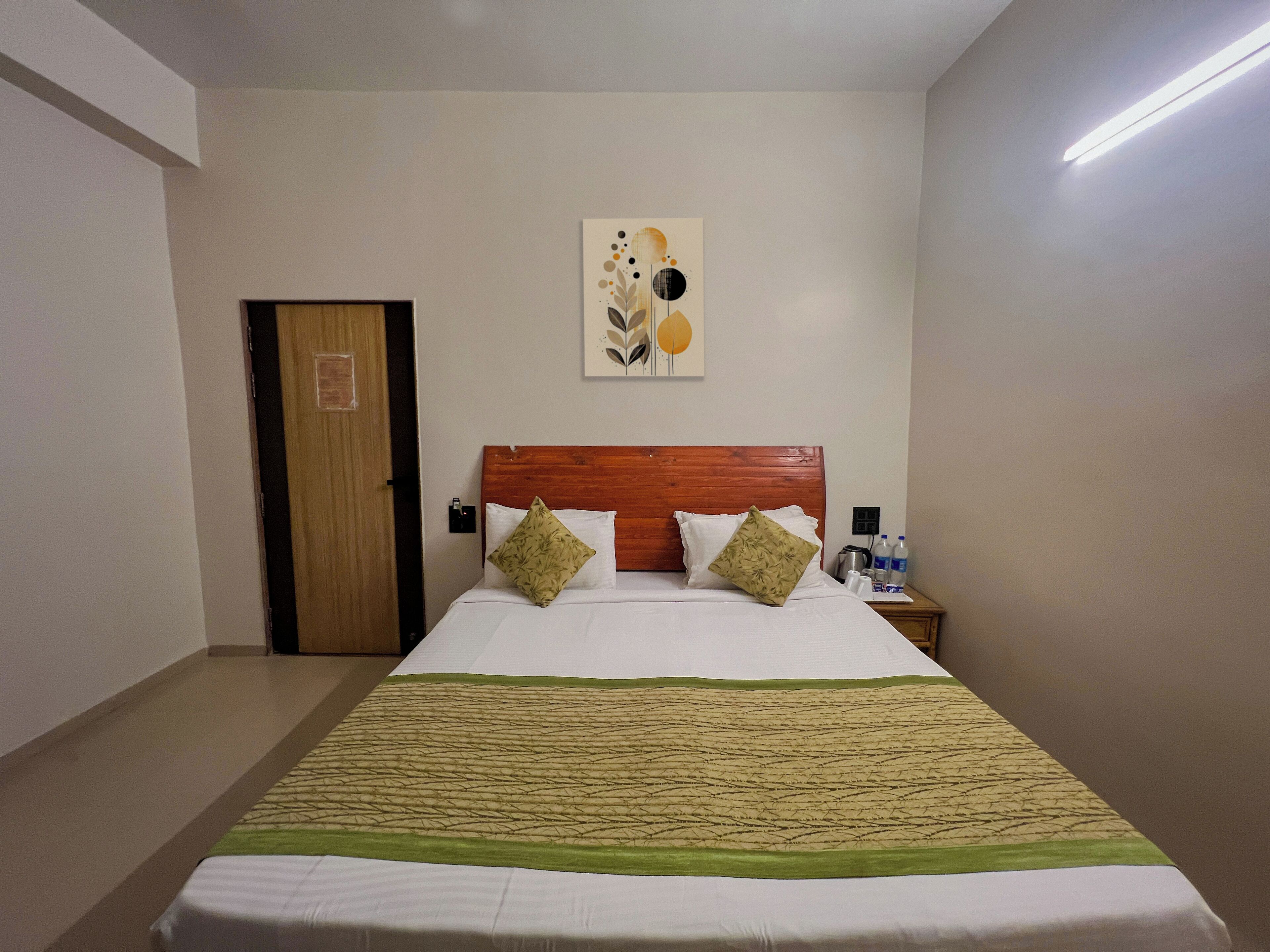 Una habitación de hotel con cabecera de madera, una cama con ropa blanca, dos almohadas decorativas, una colcha verde y beige, una obra de arte en la pared, una mesita de noche con un hervidor y una puerta.