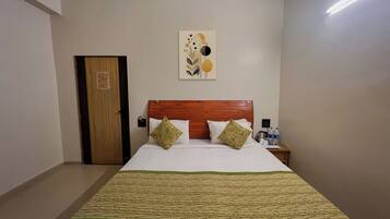 Una habitación de hotel con cabecera de madera, una cama con ropa blanca, dos almohadas decorativas, una colcha verde y beige, una obra de arte en la pared, una mesita de noche con un hervidor y una puerta.