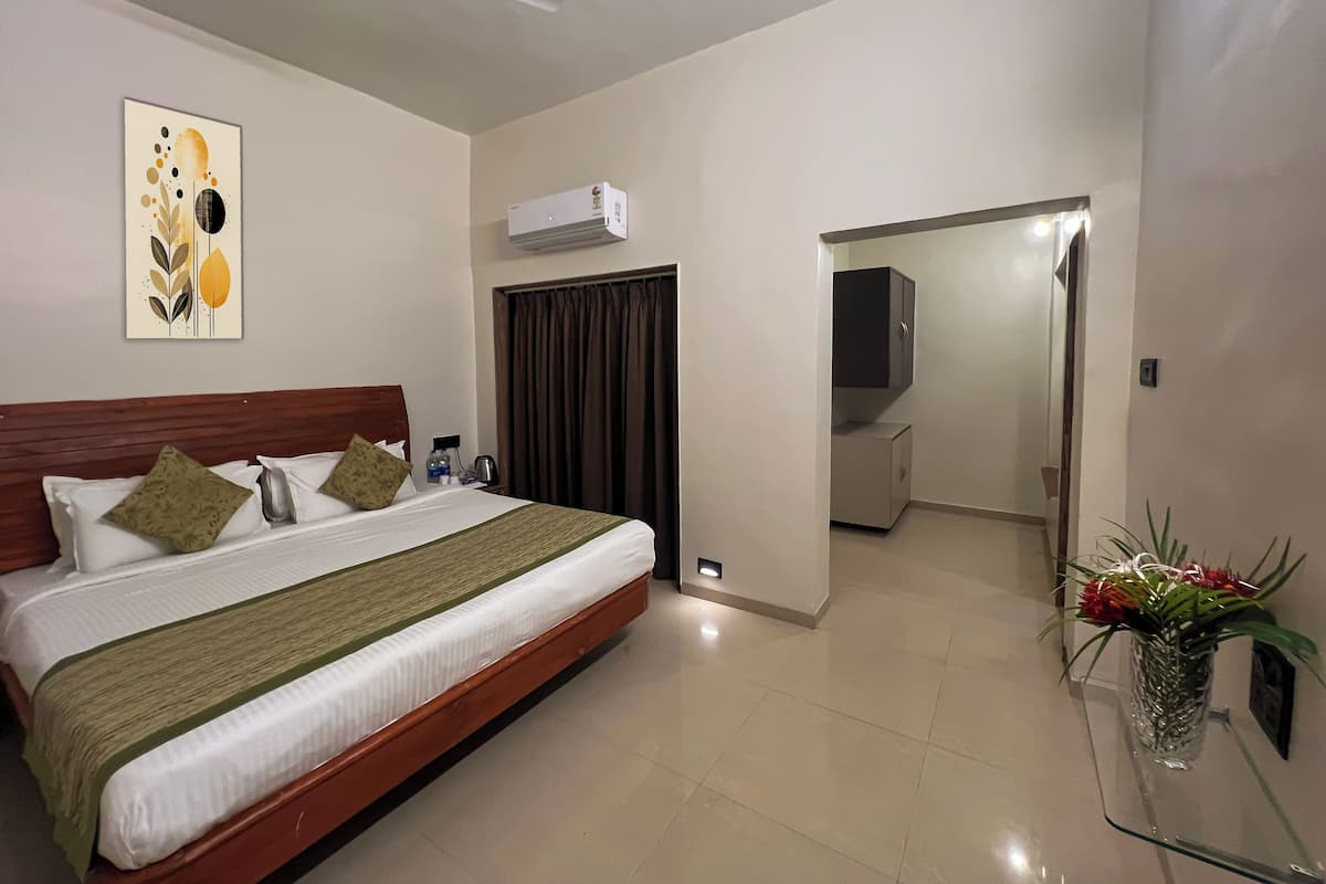 A hotel room with a bed, a ceiling fan, an air conditioner, a nightstand with a lamp, a vase with flowers, and a door leading to another room.