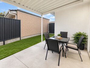 Een overdekt terras met een tafel, stoelen, een bloempot en een bakstenen muur op de achtergrond.
