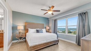 A bedroom with a bed, a ceiling fan, a nightstand with a lamp, a window with curtains, and a view of the outside. - Southern Comfort at The Aruba (Daytona Beach)