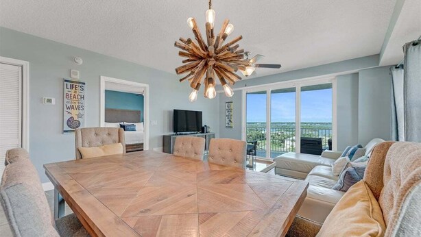 A spacious living room with a large wooden dining table, comfortable seating, a ceiling fan, and large windows offering a view of the outdoors. - Southern Comfort at The Aruba (Daytona Beach)