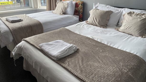 A hotel room with two beds, each with white linens and beige blankets. There is a window with curtains, a wall-mounted light fixture, and a view of the street outside. - Somerville Hotel (Blackpool)