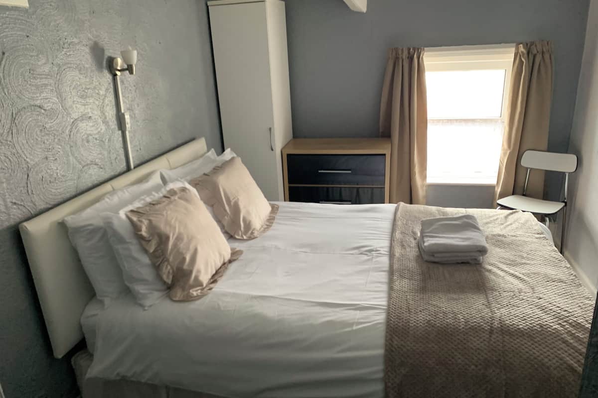 A neatly made bed with white linens and two pillows, a bedside table with a lamp, a wardrobe, and a window with curtains.