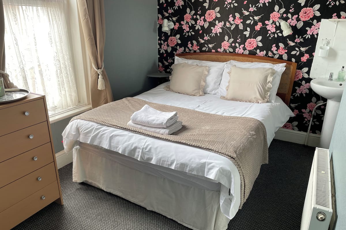 A neatly made bed with a beige blanket and pillows, a wooden dresser with drawers, a white radiator, and a floral wallpaper.