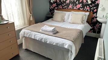 A neatly made bed with a beige blanket and pillows, a wooden dresser with drawers, a white radiator, and a floral wallpaper.