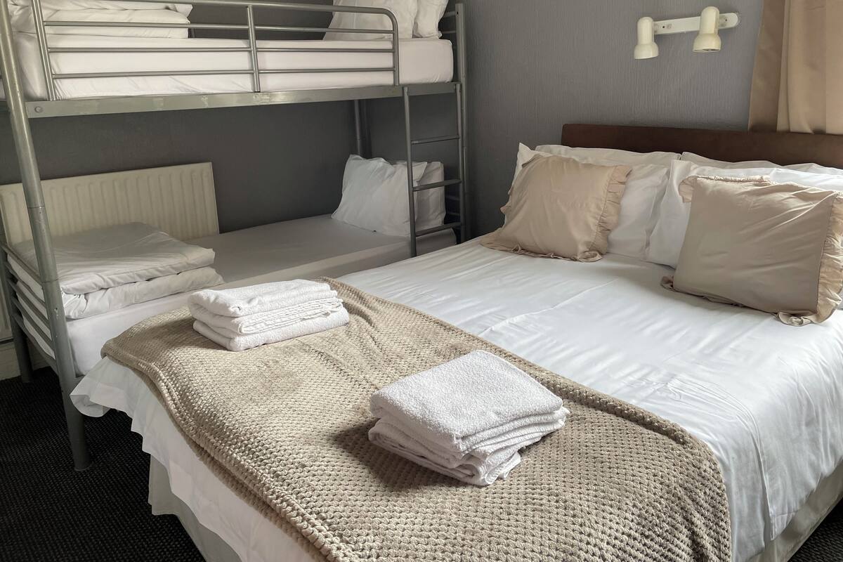 A hotel room with bunk beds, a large bed with white linens, a knitted throw blanket, and a wall-mounted light fixture.