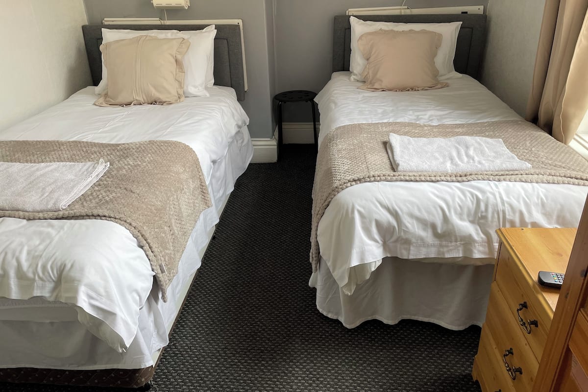 A hotel room with two single beds, each with white linens and a beige throw blanket. There is a wooden dresser with drawers and a remote control on top.