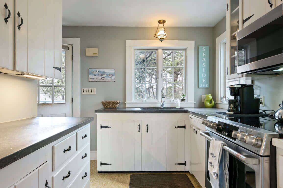 A kitchen with white cabinets, a stove, and a sink. There is a window with a view of trees and a sign that says "SEASIDE AVE".