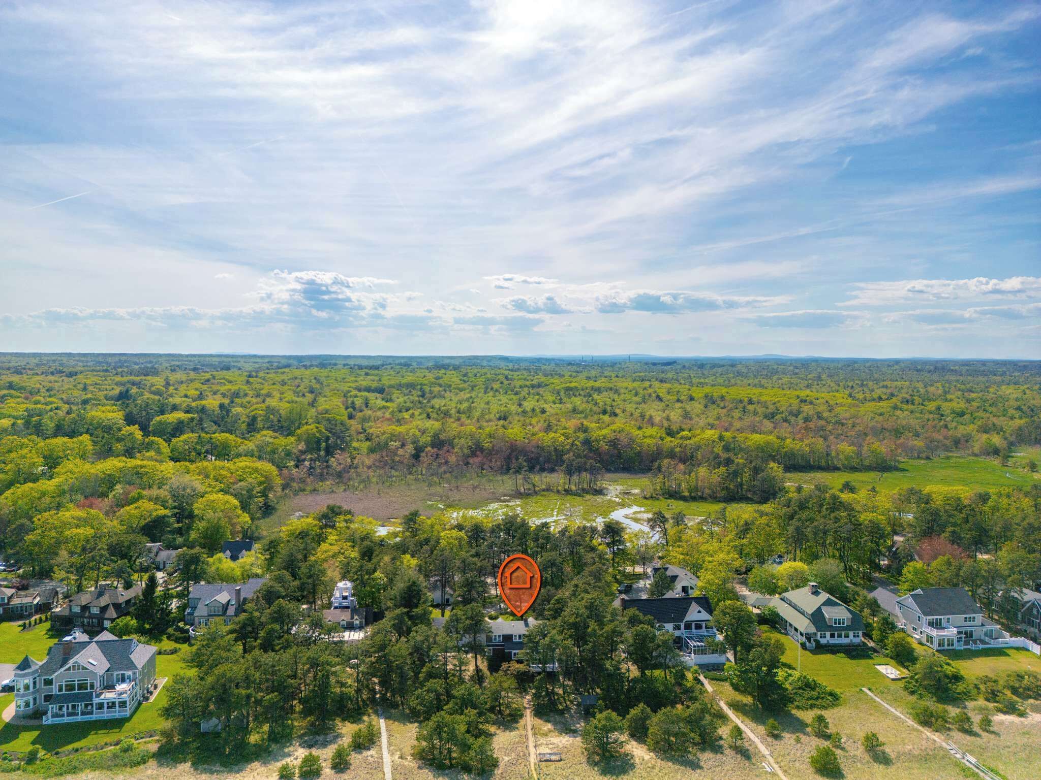 A residential area with multiple houses surrounded by dense forest, a clear sky, and a prominent location marker.