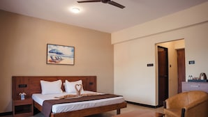 A hotel room with a large bed, a wooden headboard, a painting of a beach scene, a desk, a chair, and a ceiling fan. - Hotel Holiday Village (Alto de Porvorim)