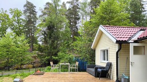 Una terraza de madera con mobiliario exterior, una casita con techo de tejas rojas y un bosque al fondo.