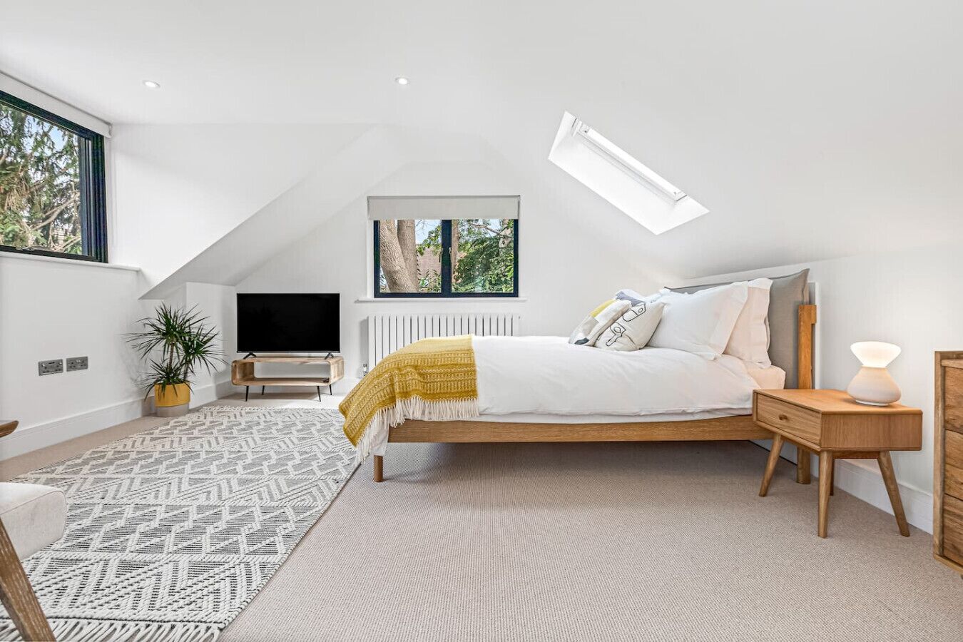 A modern bedroom with a sloped ceiling, a bed with a wooden frame, a bedside table with a lamp, a TV, and a large window with a view of trees.