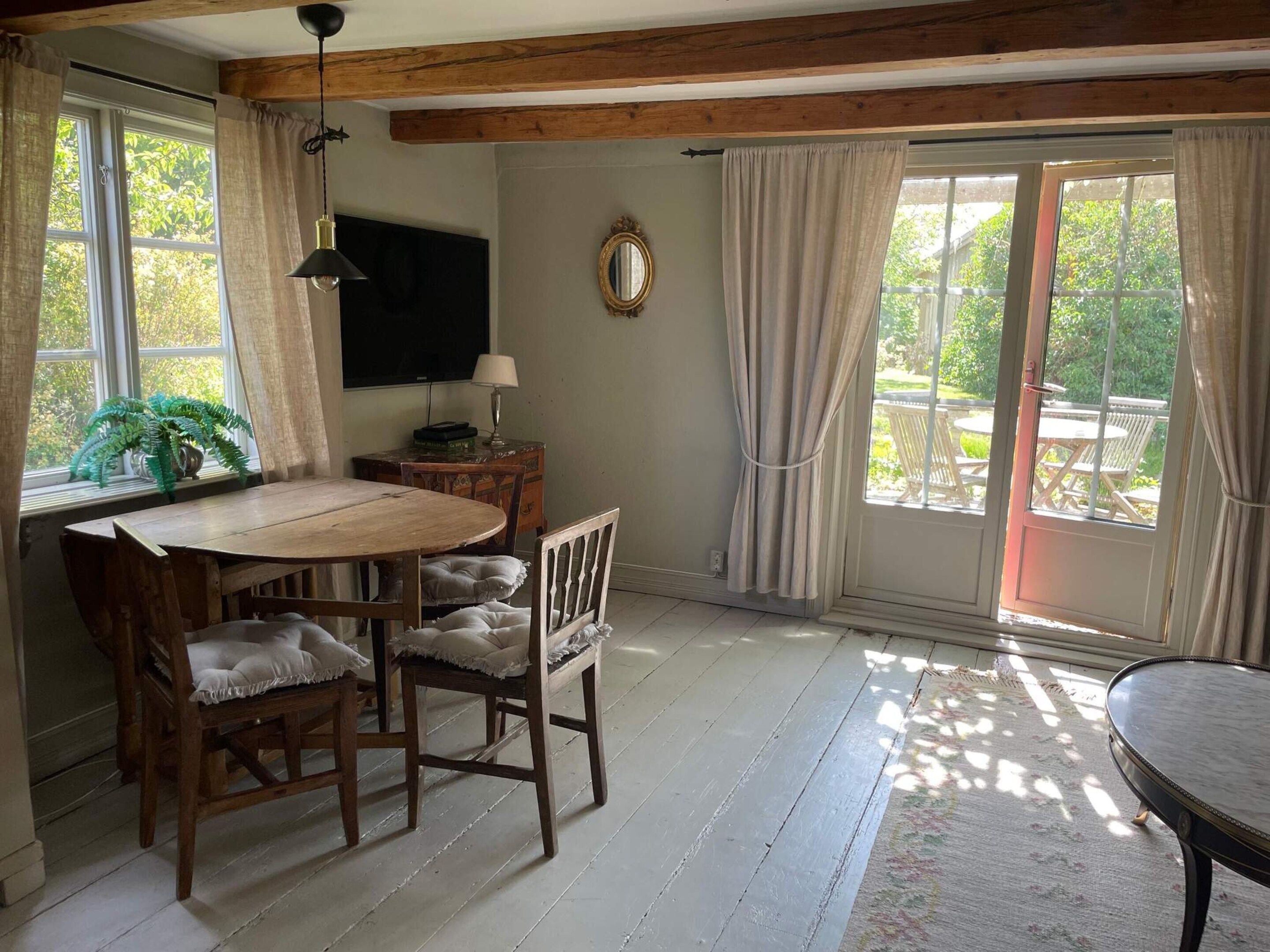 Un espace repas comprenant une table et des chaises en bois, un téléviseur, un miroir et une vue sur l’extérieur grâce à de grandes fenêtres.