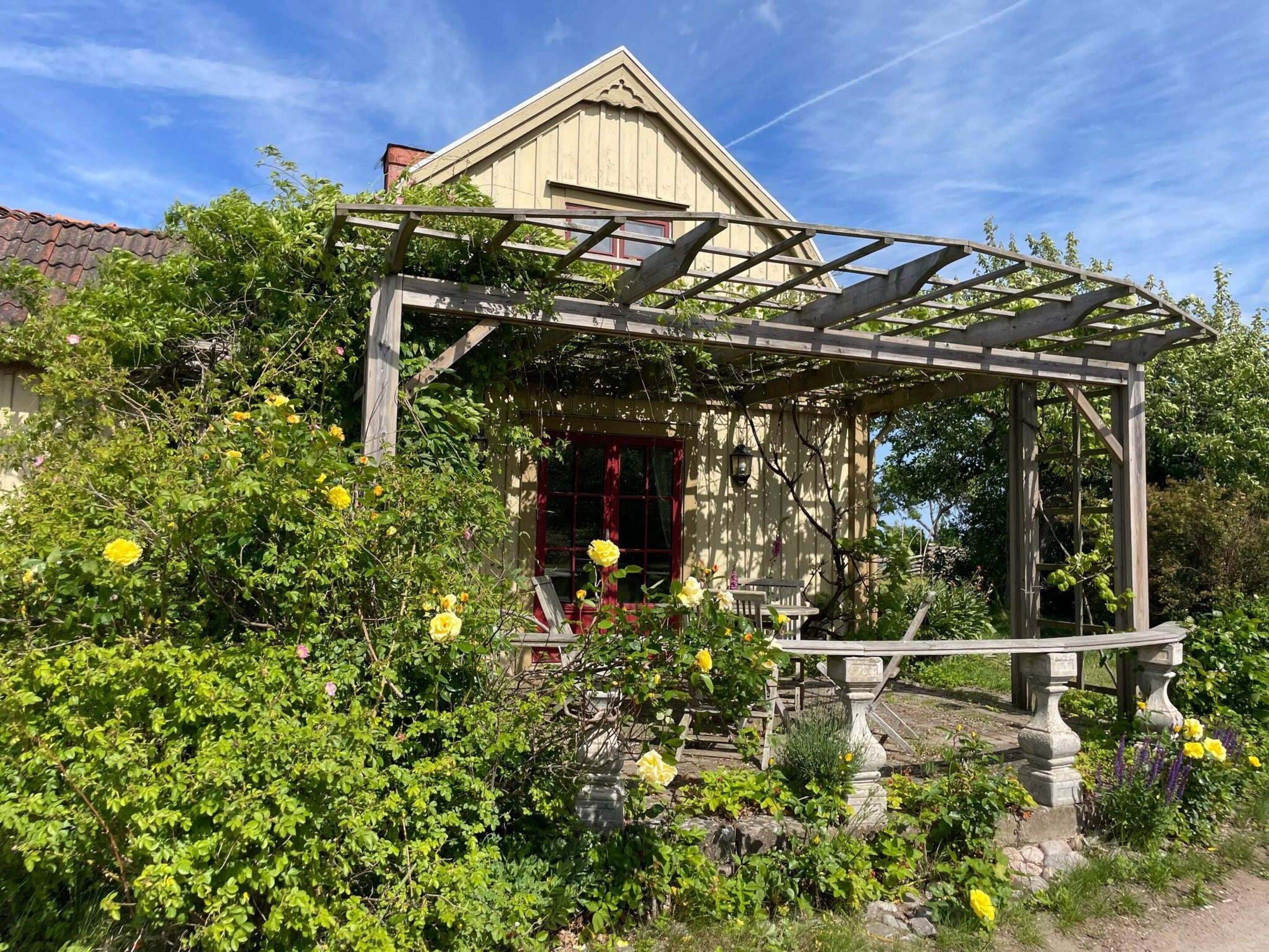 A wooden pergola with a stone bench, surrounded by lush greenery and yellow flowers.