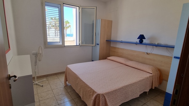 Een kleine, schone kamer met een bed, een raam, een spiegel, een ventilator en een plank met een lamp.