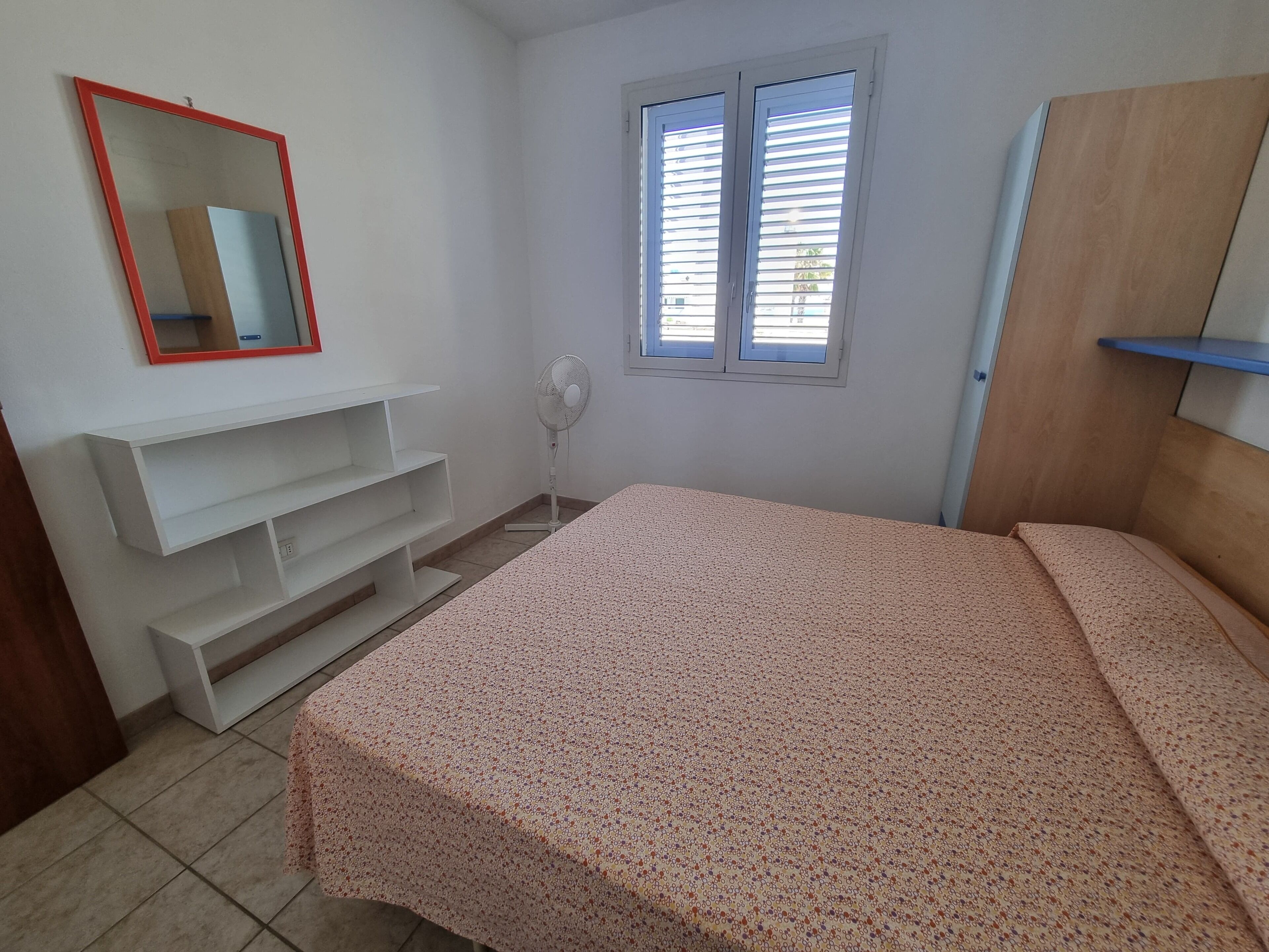 A bedroom with a bed, a white TV stand, a mirror, a window with blinds, and a fan.