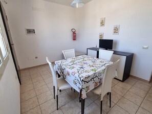 Un coin repas avec une table recouverte d’une nappe ornée de motifs d’arbres et de hiboux, des chaises blanches, un petit bureau noir et un extincteur fixé au mur.