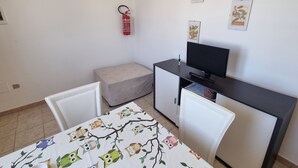 Een kleine, schone kamer met een bed, een bureau met een computer, een televisie en een brandblusser aan de muur.