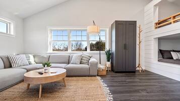 A modern living room with a grey sofa, a round wooden coffee table, a grey cabinet, and a large window with natural light.