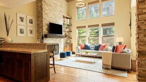 A spacious living area with a stone fireplace, a television mounted above it, a sofa with colorful cushions, and a large window offering a view of greenery.