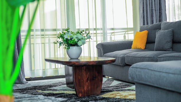 Ein Wohnzimmer mit grauem Sofa, einem Holztisch mit weißer Blumenvase und einem großen Fenster mit durchscheinenden Vorhängen.