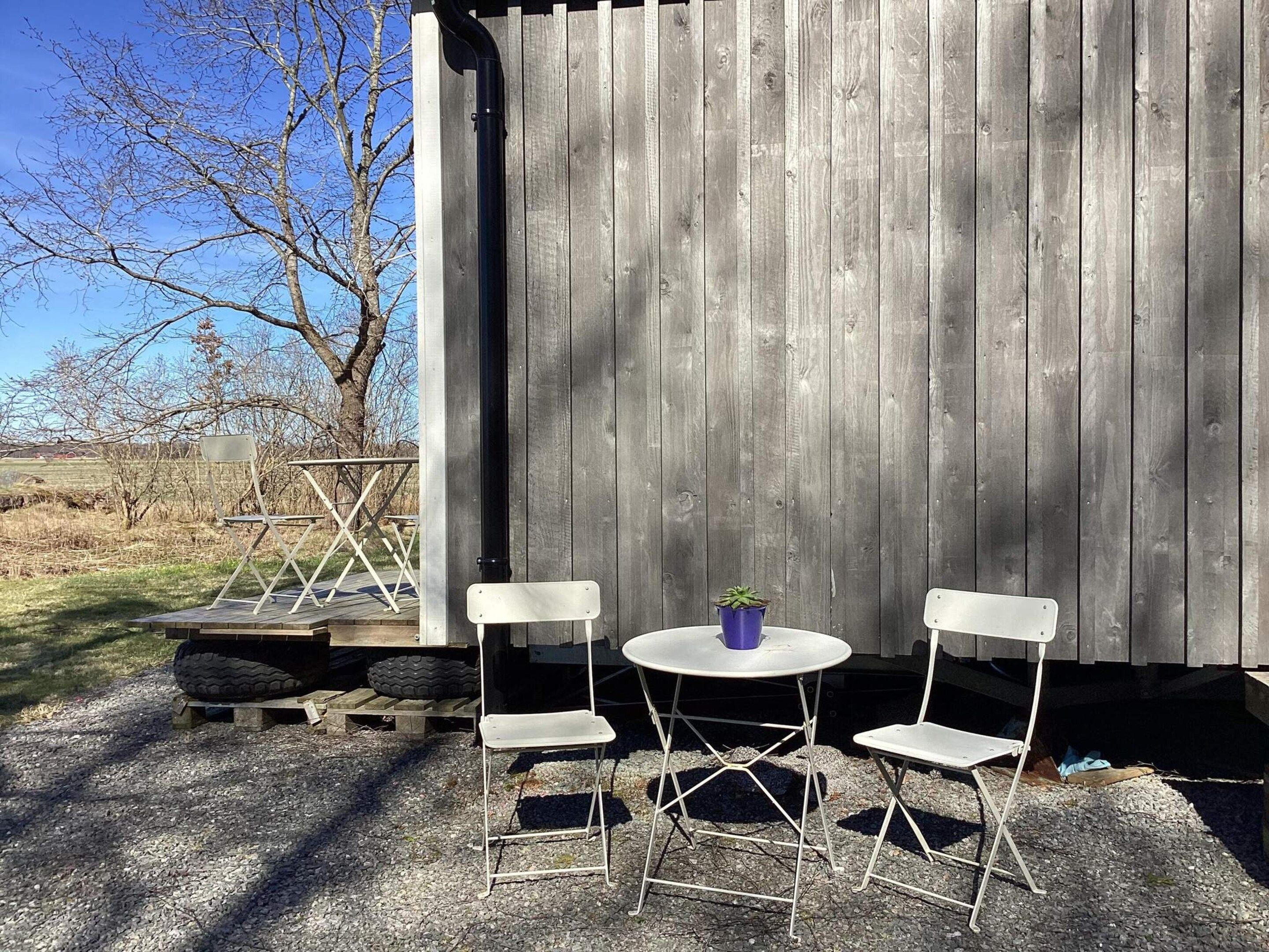 A rustic outdoor seating area with a wooden table and two chairs, a potted plant, and a tire under a wooden structure.