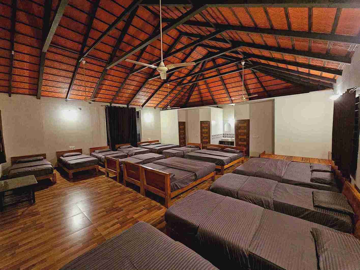 A dormitory-style room with multiple beds arranged in rows, wooden floors, and a high, vaulted ceiling with exposed beams.