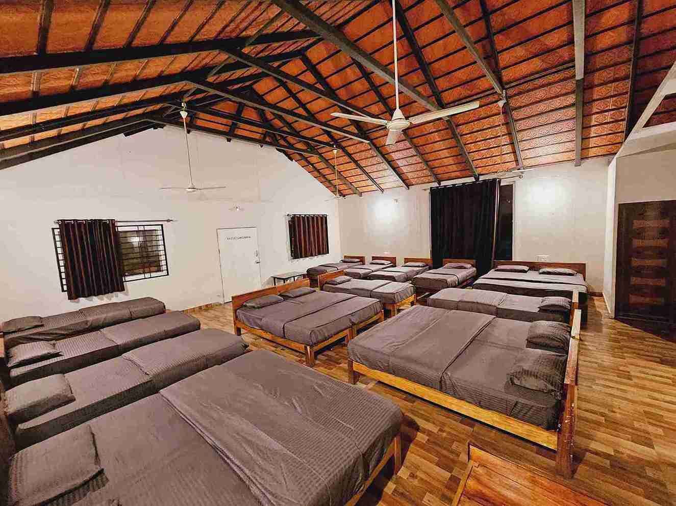 Una habitació de dormitori amb llits múltiples disposats per files, terres de fusta i un sostre inclinat amb bigues al descobert.