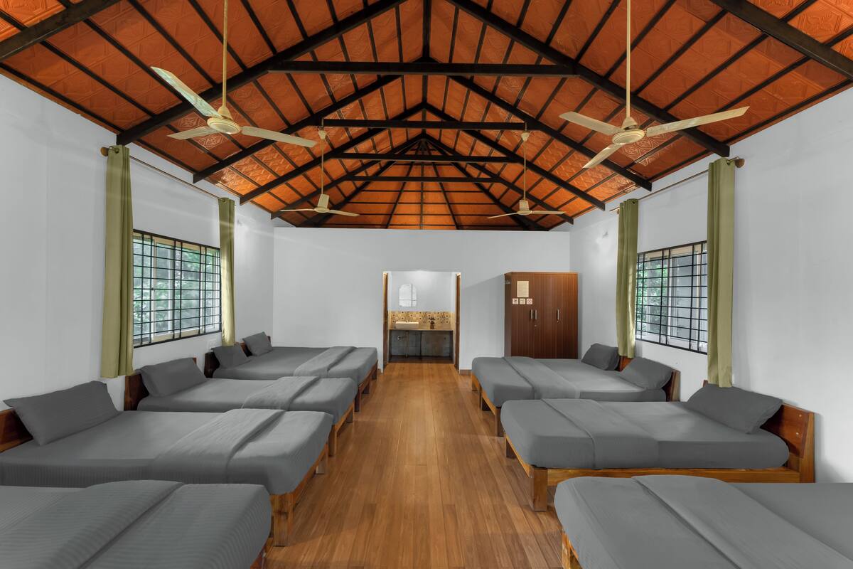 A room with multiple beds arranged in rows, a wooden ceiling, and a small kitchenette in the center.