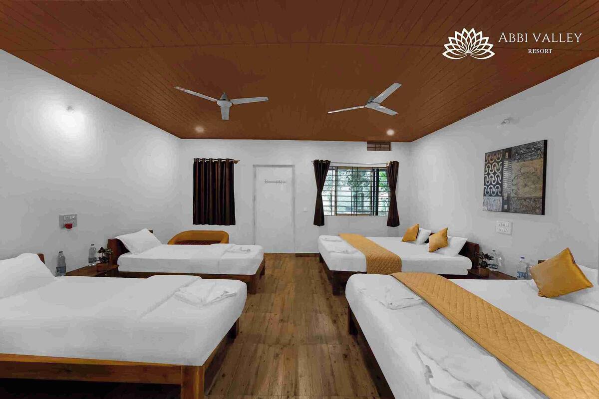 A hotel room with four single beds, wooden flooring, ceiling fans, a window with curtains, and a wall-mounted artwork.