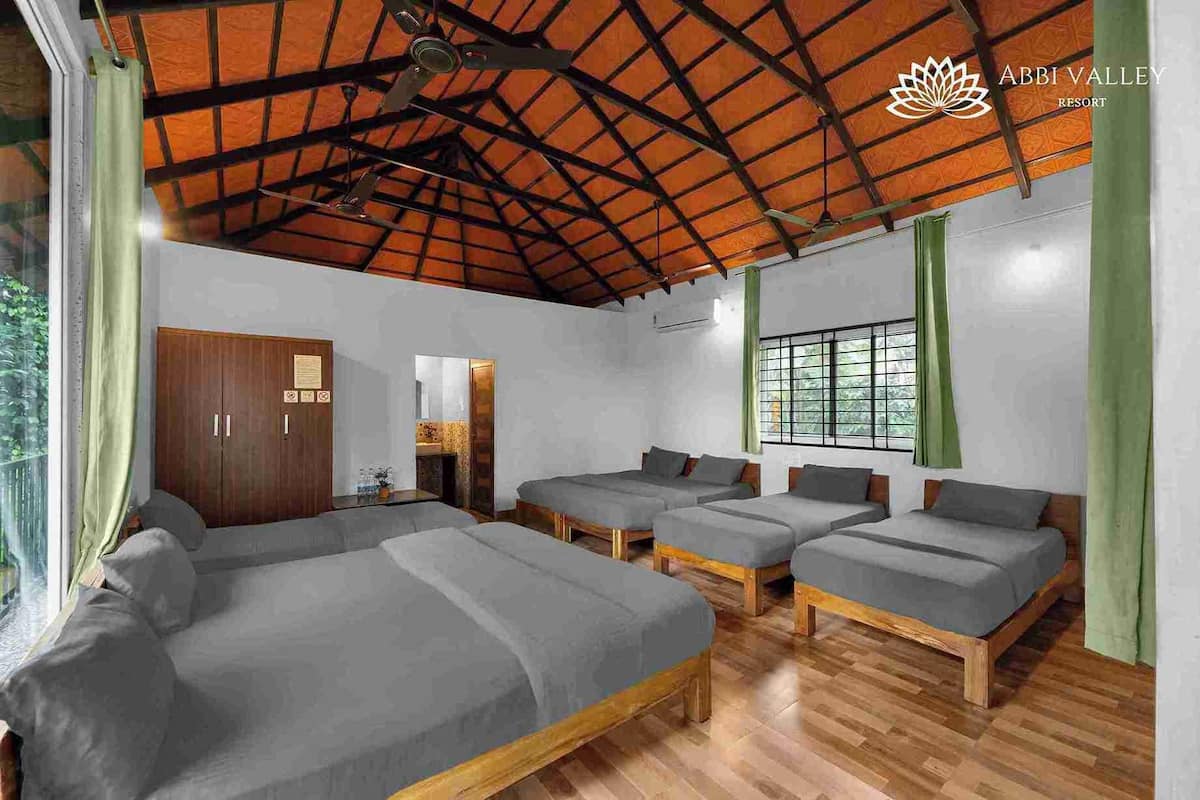 A room with four beds, a wooden wardrobe, a window with green curtains, and a ceiling with a wooden beam structure.
