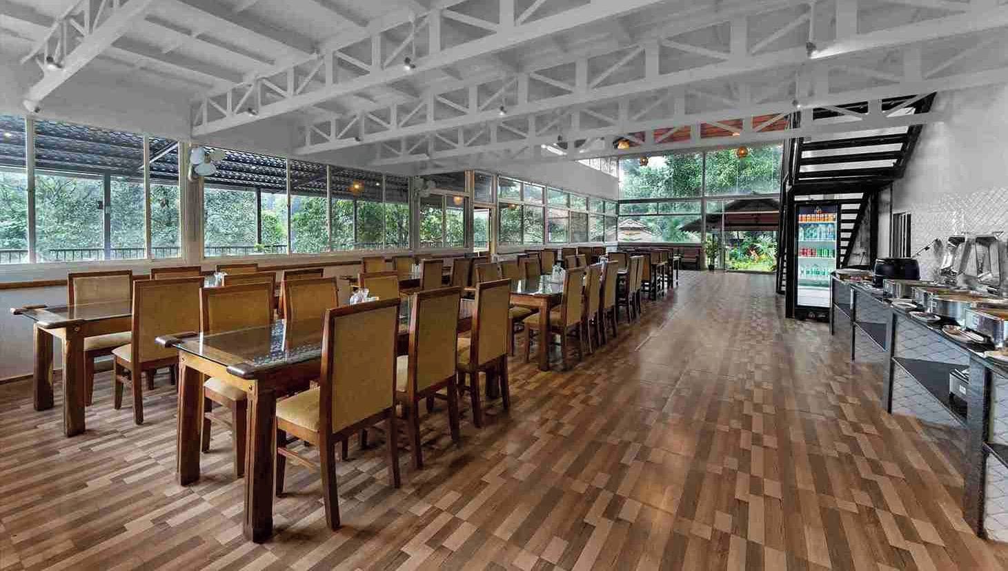 A spacious dining area with wooden tables and chairs, large windows offering views of greenery, and a buffet setup on the right.