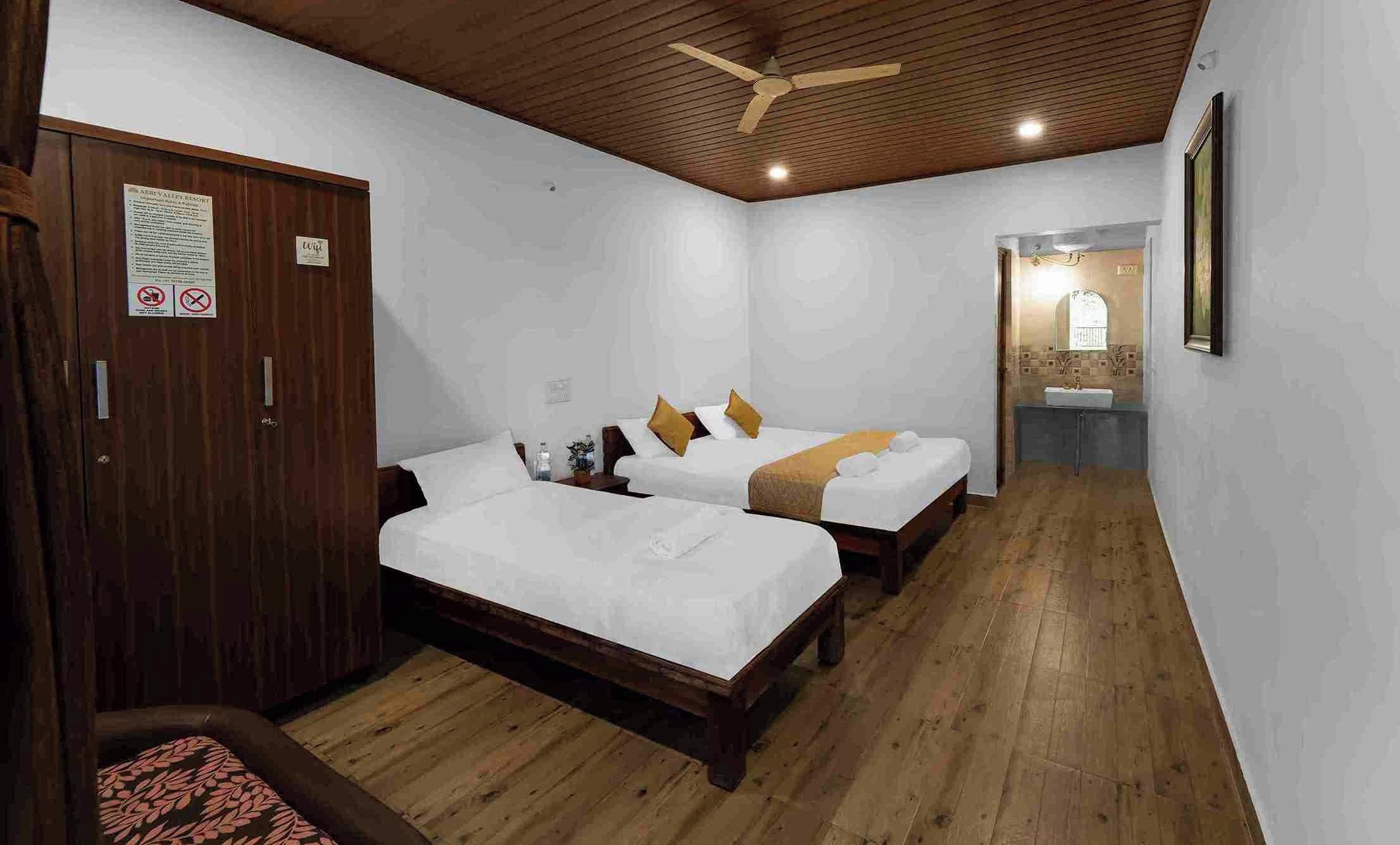 Habitación de hotel con dos camas, piso de madera, ventilador de techo y baño con lavamanos y espejo.