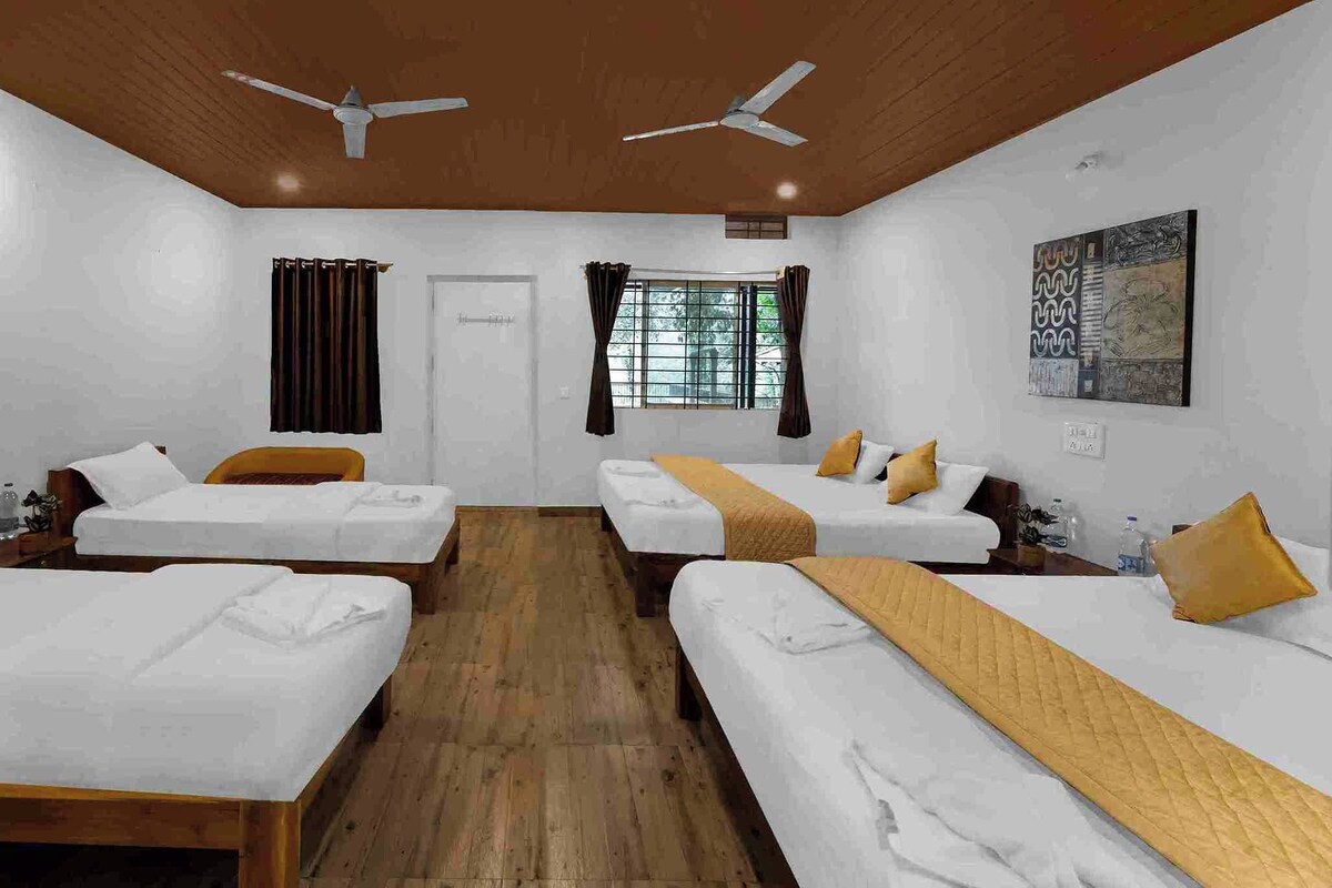 A hotel room with four single beds, wooden flooring, ceiling fans, a window with curtains, and a wall-mounted artwork.