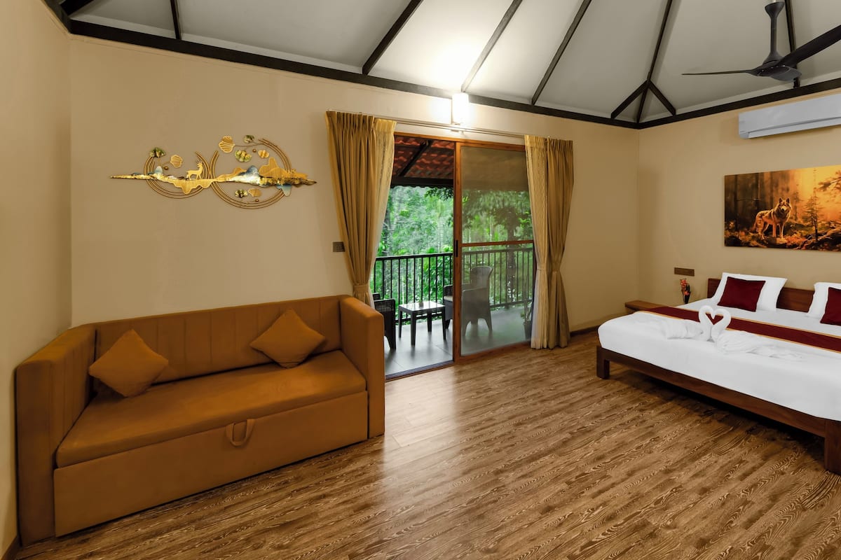 A hotel room with a wooden floor, a bed with white bedding and red pillows, a brown sofa, a ceiling fan, and a painting on the wall.