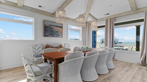 A dining area with a wooden table and white upholstered chairs, large windows with a view of the sea, and a hanging light fixture. - Experience luxury at SeaSons at the Point, a stunning oceanfront rental in Oak Island. (Oak Island)