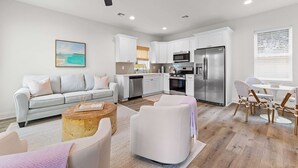 A modern living room with a sofa, coffee table, and chairs, a kitchen with white cabinets and appliances, and a ceiling fan. - New Breezy Cottage - King Bed - Mini-Golf - Park (Youngsville)