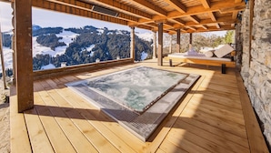 Een houten terras met een jacuzzi en een bank, met uitzicht op een besneeuwd berglandschap.