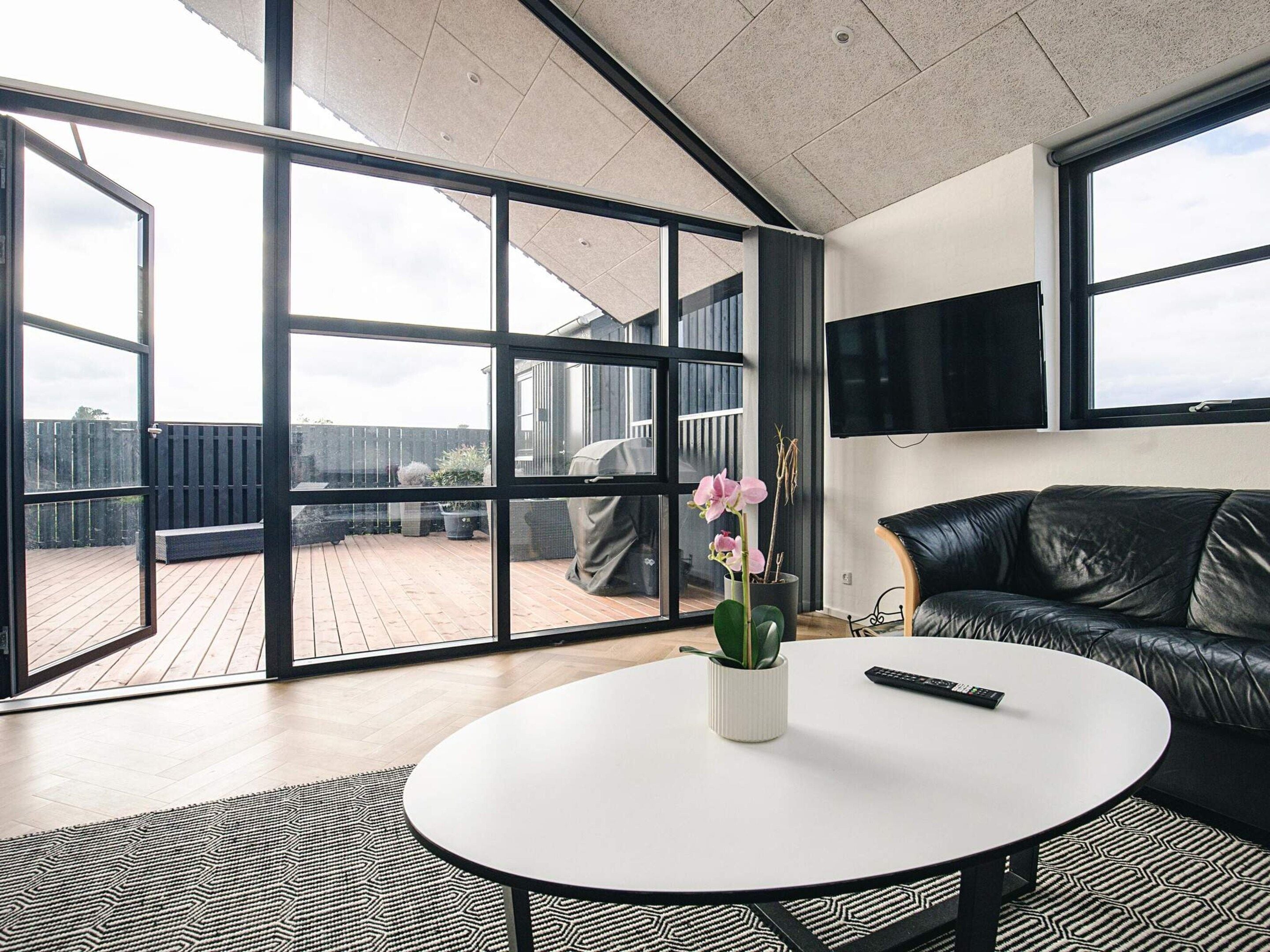 A modern living room with a large glass door leading to a wooden deck, a round white table, a black leather sofa, and a mounted TV.