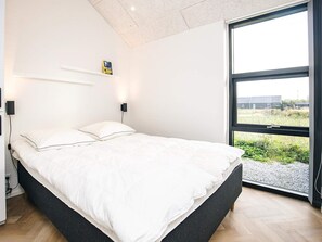 A modern bedroom with a large bed, bedside lamps, a small shelf with a book, and a view of the outdoors through a large window. - 5 Star Holiday Home in Lokken (Lokken)