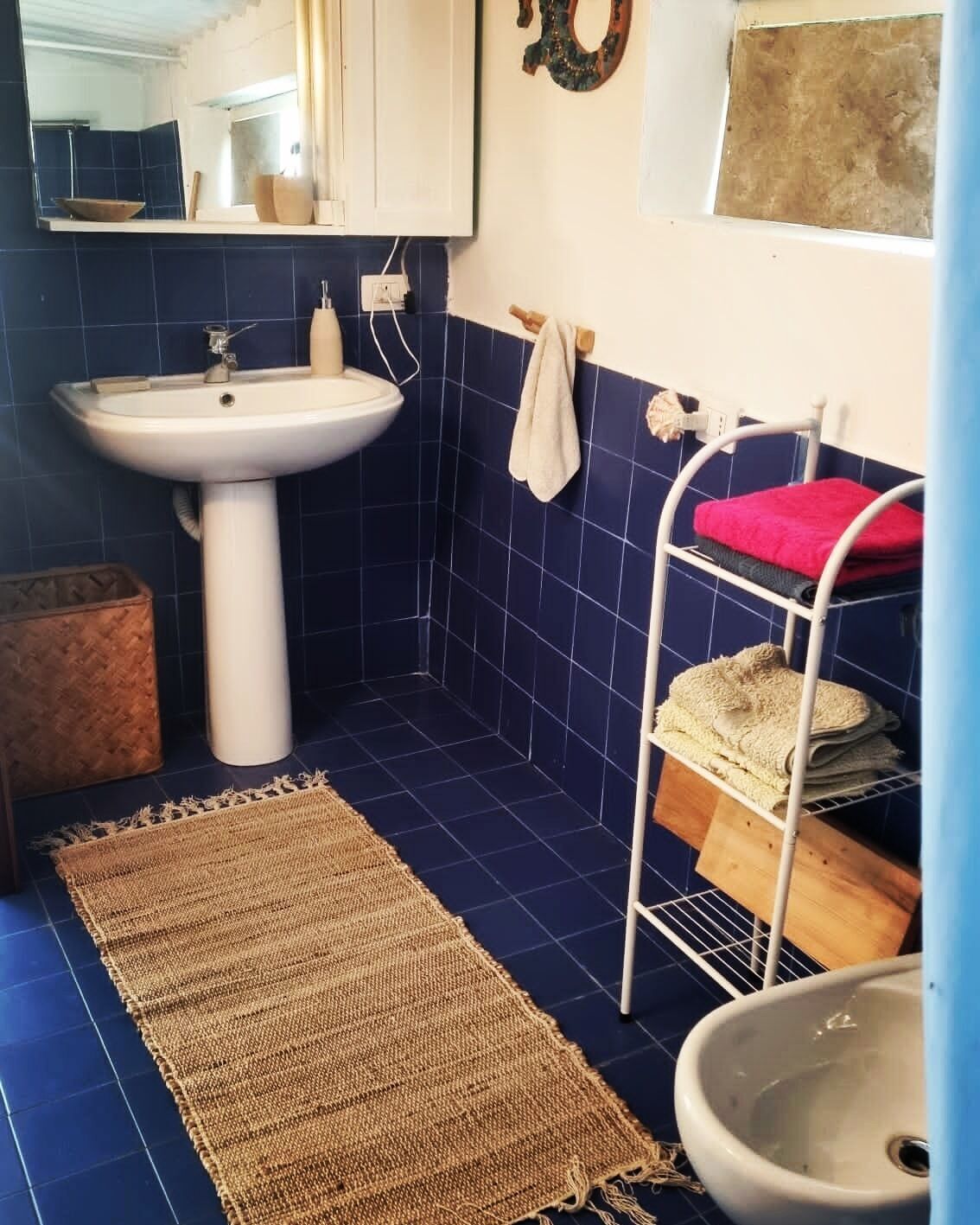 A bathroom with a white sink, a mirror, a towel rack with a red towel, a shelf with towels, and a small trash bin.